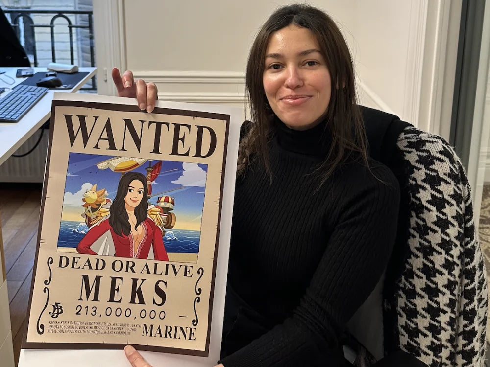 Une femme assise dans un bureau sourit en tenant une affiche Wanted One Piece personnalisée d'elle-même dans un style pirate. L’illustration inclut le Thousand Sunny en arrière-plan, une prime affichée et des ornements rappelant les avis de recherche de l'anime One Piece