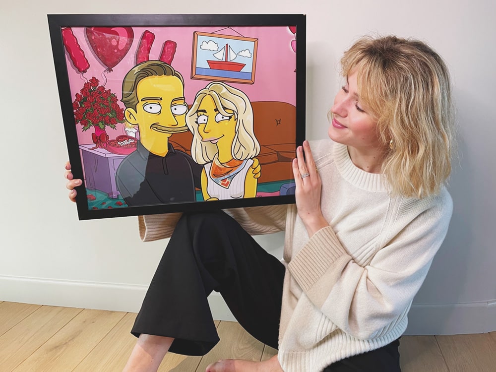 Une femme assise sur le sol, souriant tout en tenant un portrait de couple encadré et personnalisé de style des Simpson. L'illustration présente les personnages dessinés dans le salon des Simpson, avec des ballons et des fleurs